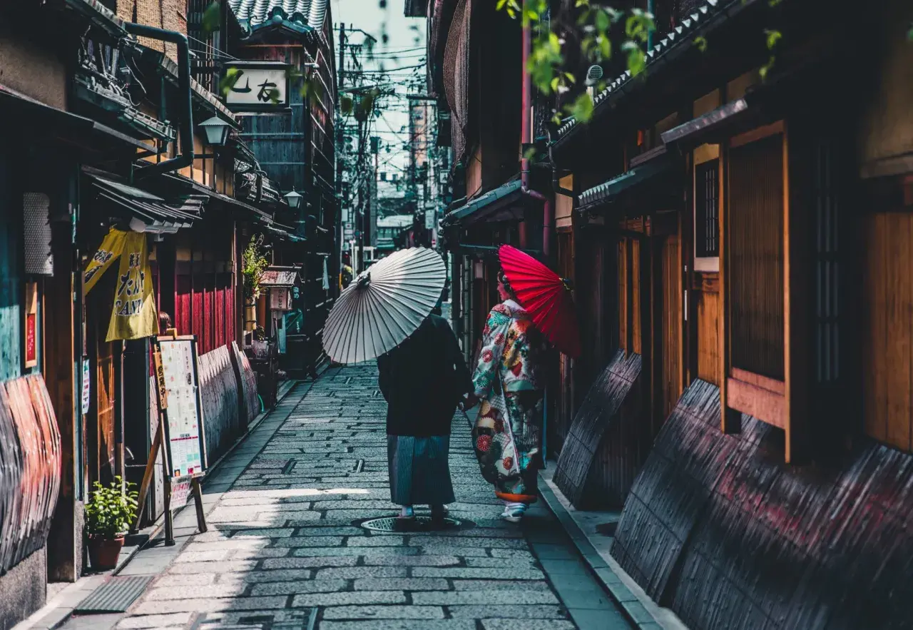 Dwie kobiety w kimonach spacerują wąską uliczką w Kioto, trzymając parasolki. To fascynujące ciekawostki o Japonii.