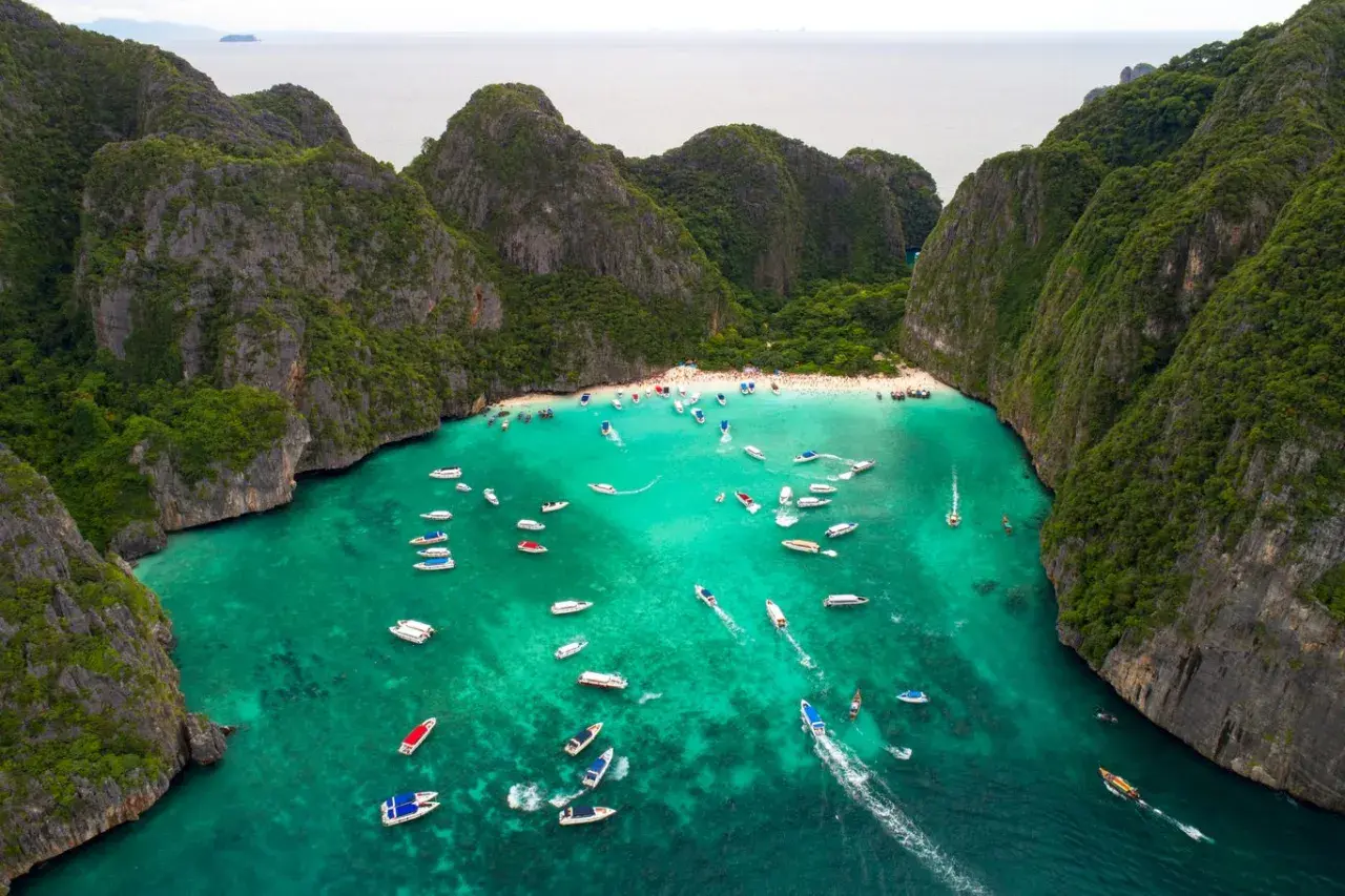 Maya Bay tętni życiem, pełna łodzi i turystów na piaszczystej plaży, otoczona stromymi, zielonymi klifami.