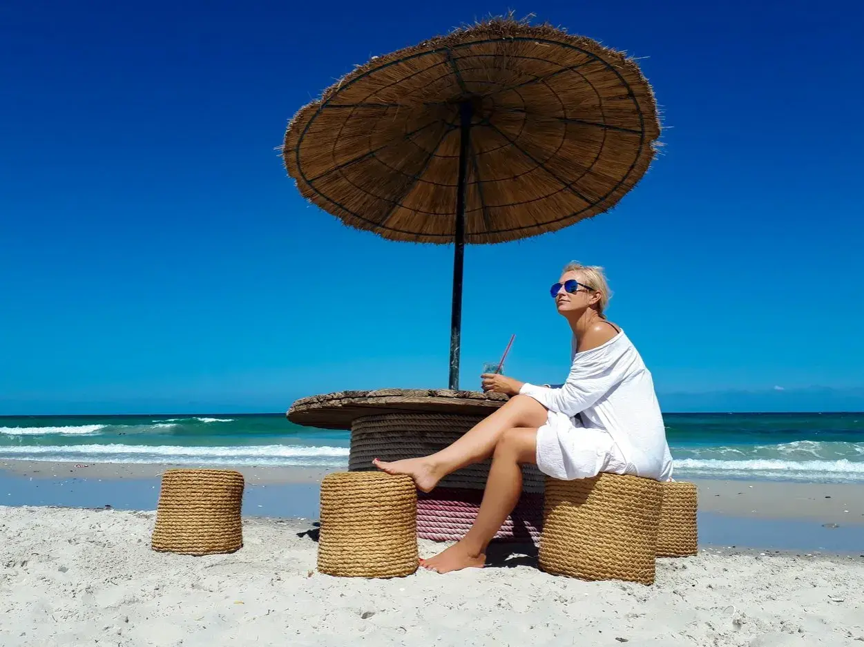Kobieta siedzi pod parasolem na plaży, popijając napój. Ceny w Tunezji są przystępne, co pozwala na takie relaksy.
