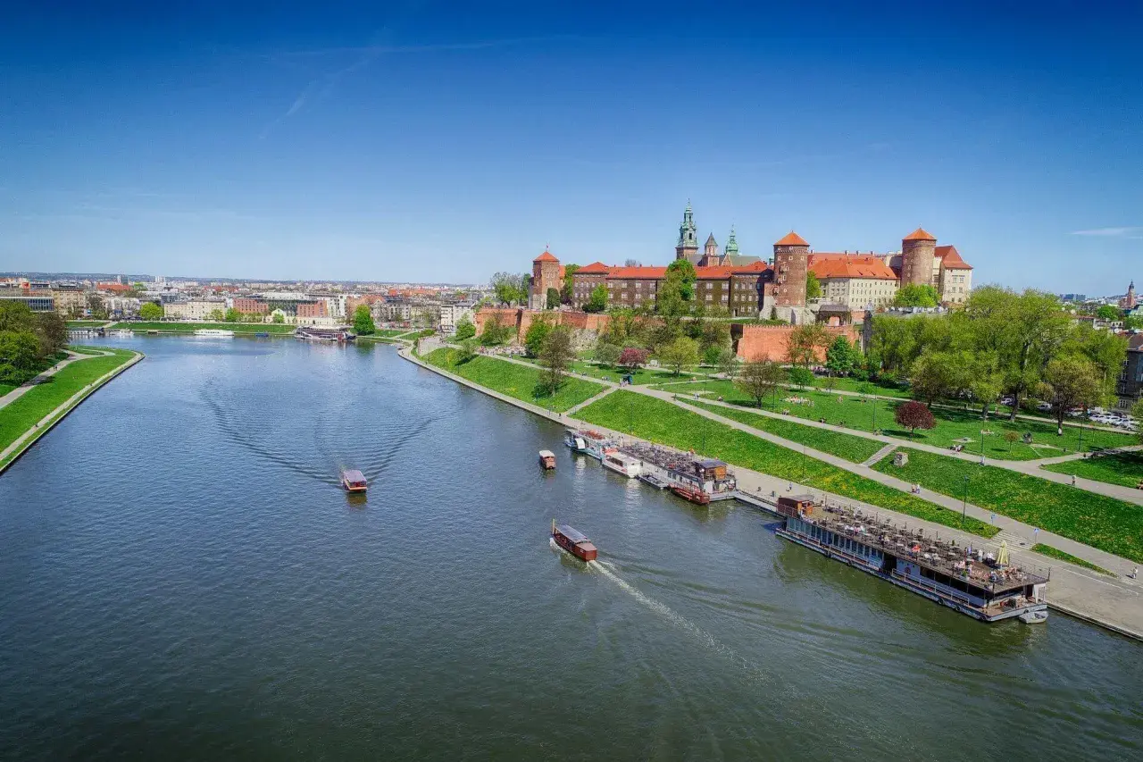 Widok na Wawel i rzekę Wisłę z łodziami. Słoneczny dzień zachęca do spacerów po zielonych bulwarach.