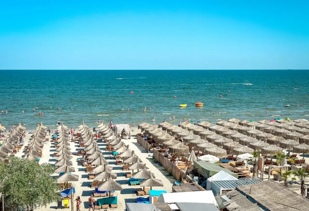Słoneczny dzień na plaży nad Morzem Czarnym w Rumunii. Rzędy parasoli, ludzie kąpiący się i pływający.
