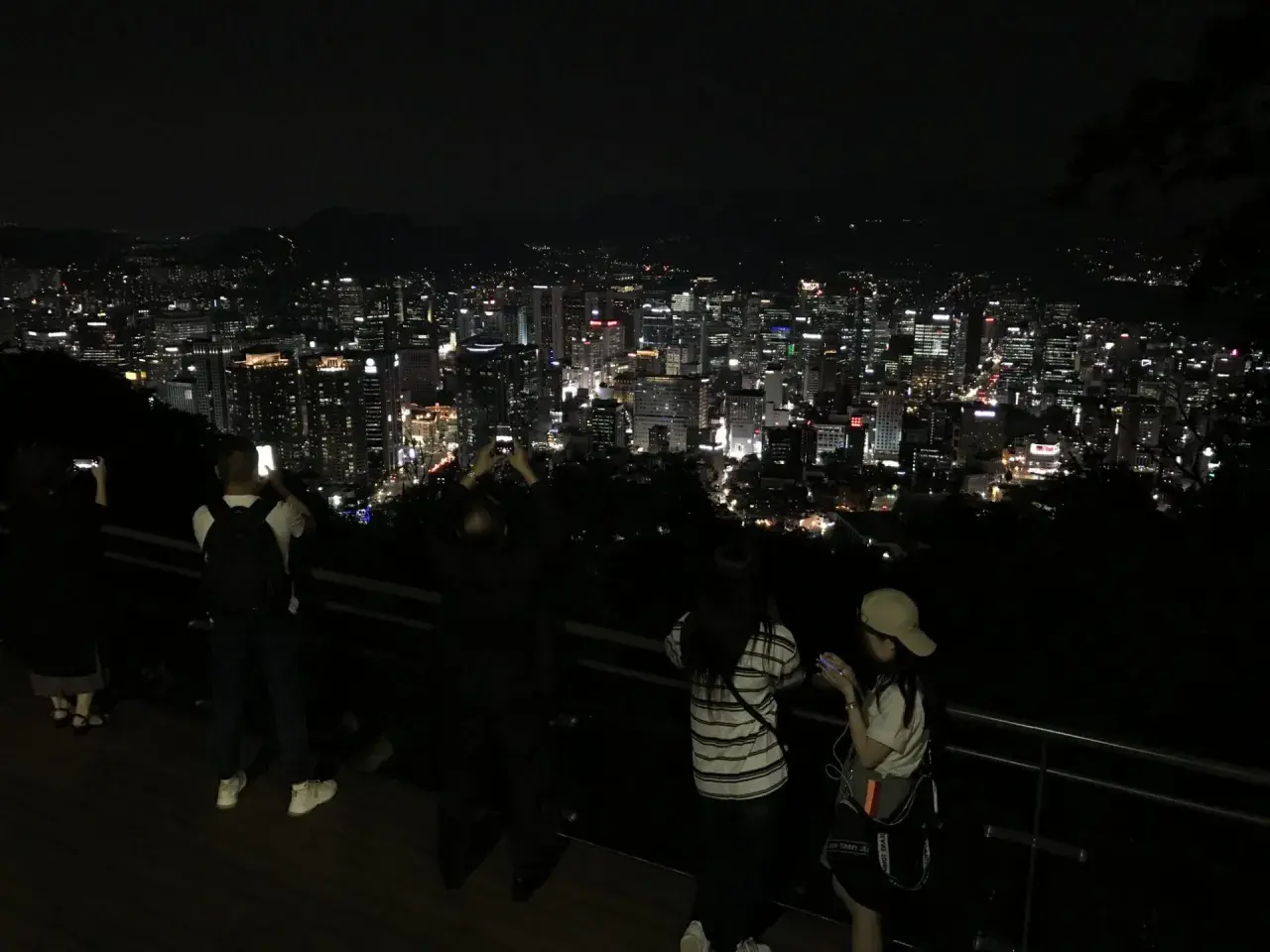 Nocny widok na rozświetlone miasto, przypominające panoramę Gangnam. Ludzie robią zdjęcia, podziwiając miejski krajobraz.