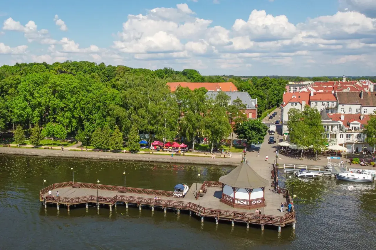 Drewniany pomost z altaną nad jeziorem, zielone drzewa i miasteczko z czerwonymi dachami. Idealne miejsce, gdzie na Mazury.
