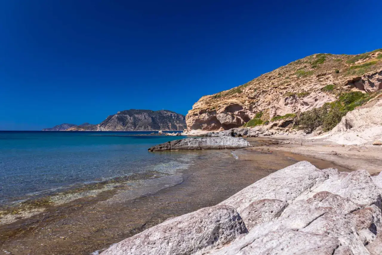 Majowa pogoda na Kos: skalista plaża z turkusową wodą i łagodnymi falami, w tle góry.
