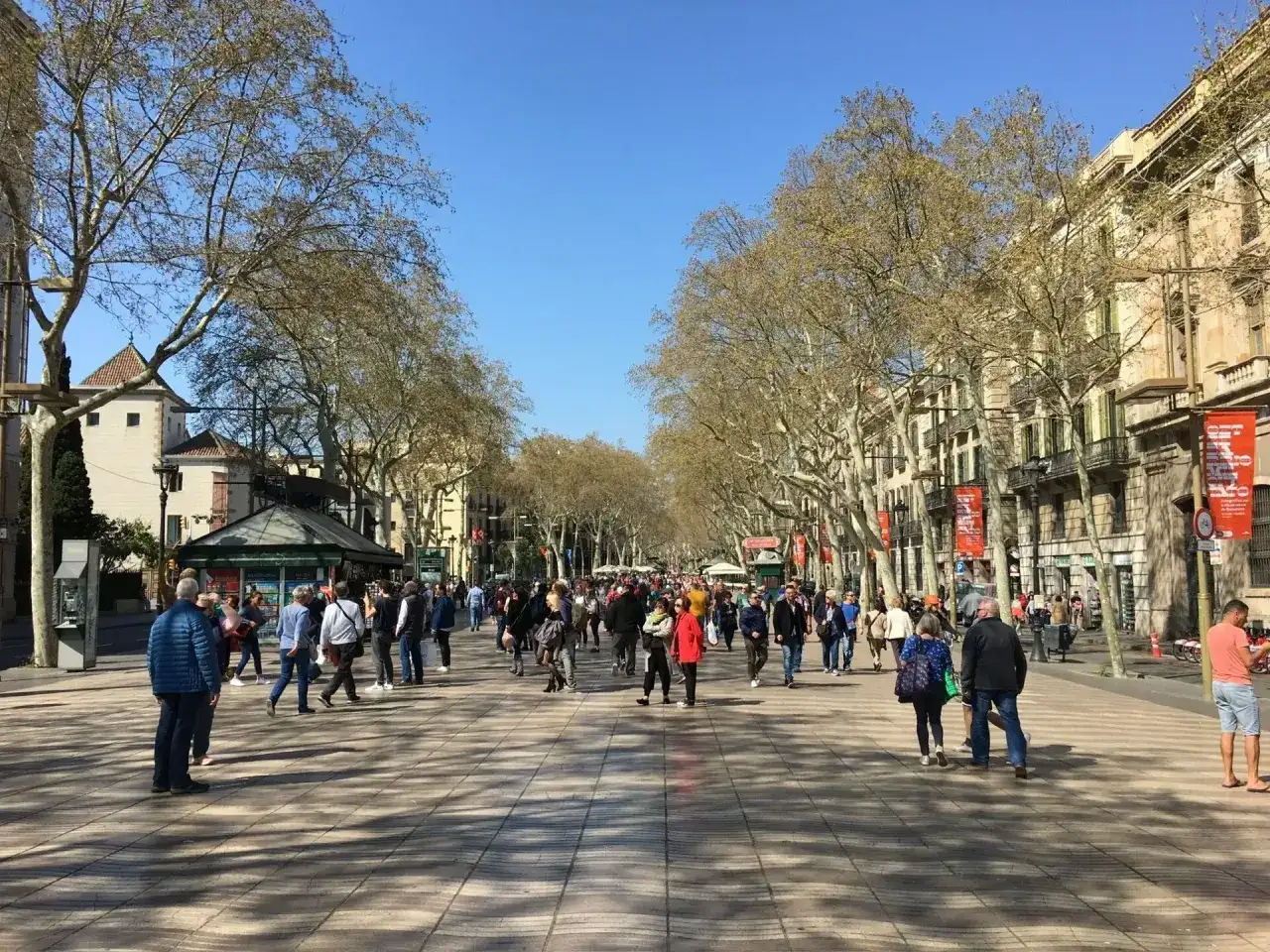 Tłum ludzi spaceruje po słonecznej La Rambla w Barcelonie, otoczonej drzewami i zabytkowymi budynkami.