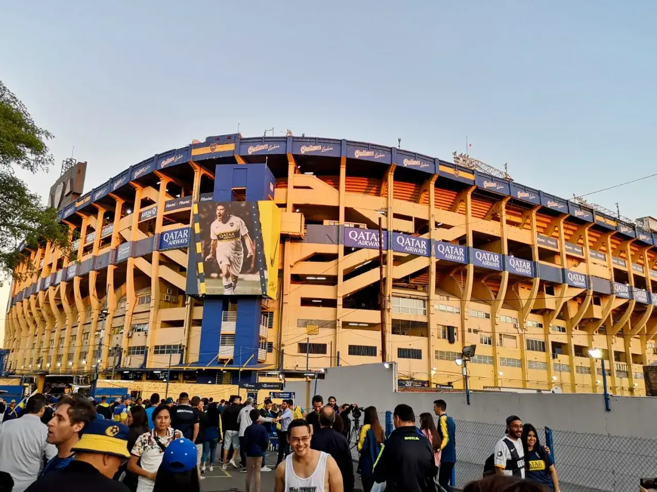 Tłum kibiców przed stadionem La Bombonera. Wielki baner z piłkarzem zdobi żółto-niebieską fasadę.