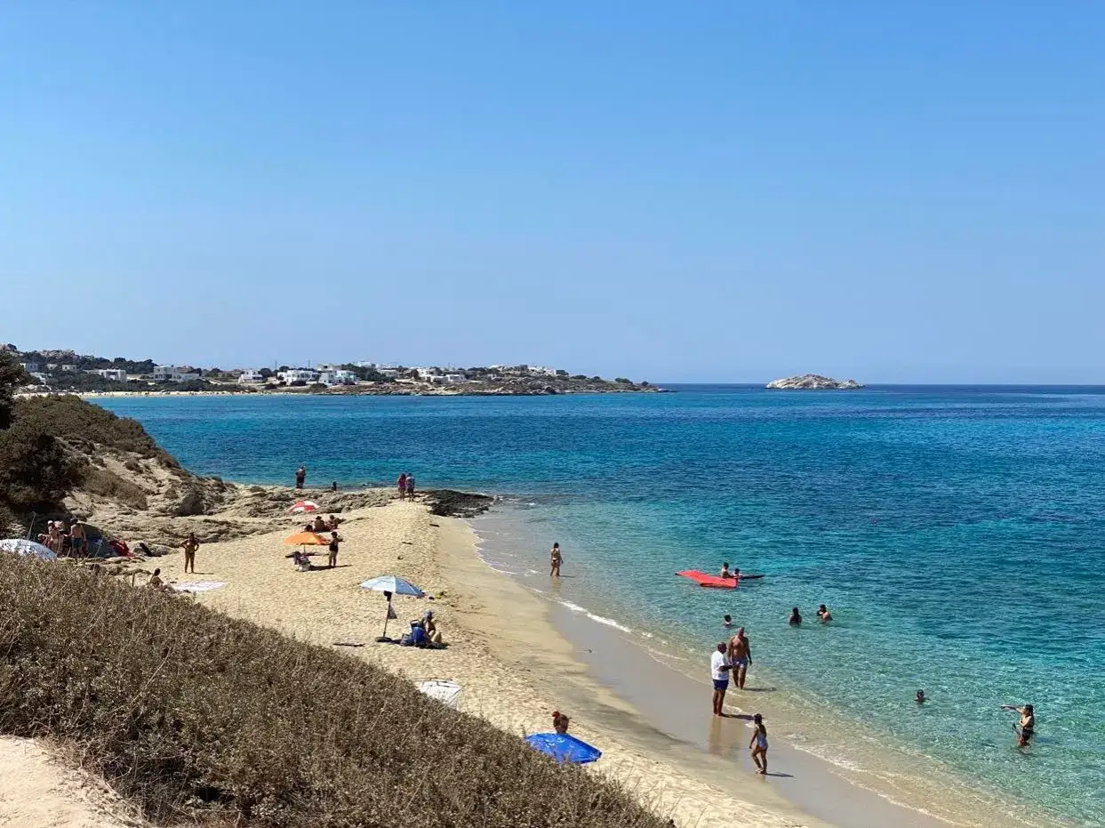 Słoneczna plaża na Naxos z turkusową wodą, białymi domkami w oddali i ludźmi relaksującymi się na piasku.