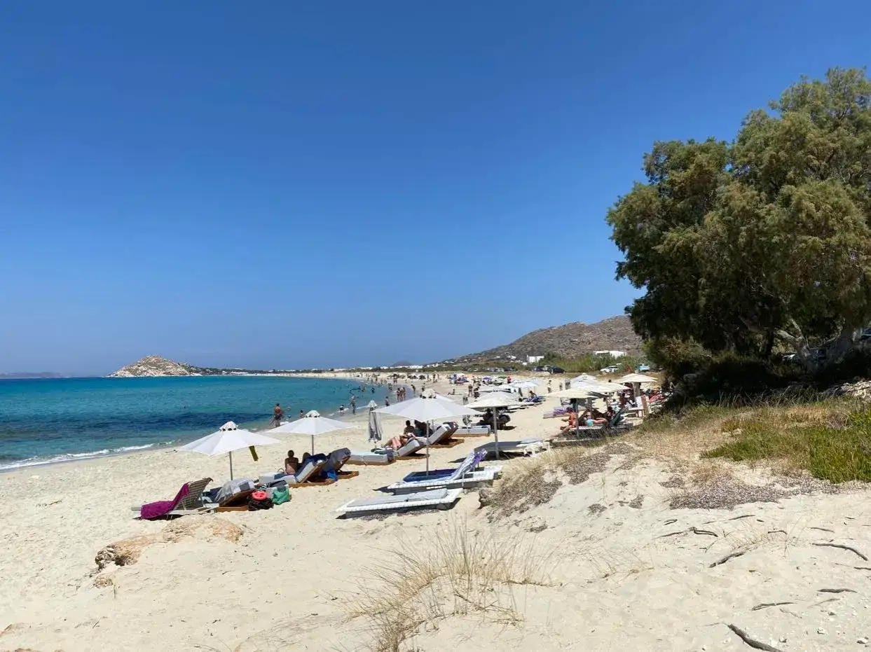 Słoneczna plaża na Naxos z turkusową wodą, białymi parasolami i leżakami. W tle wzgórza i morze.
