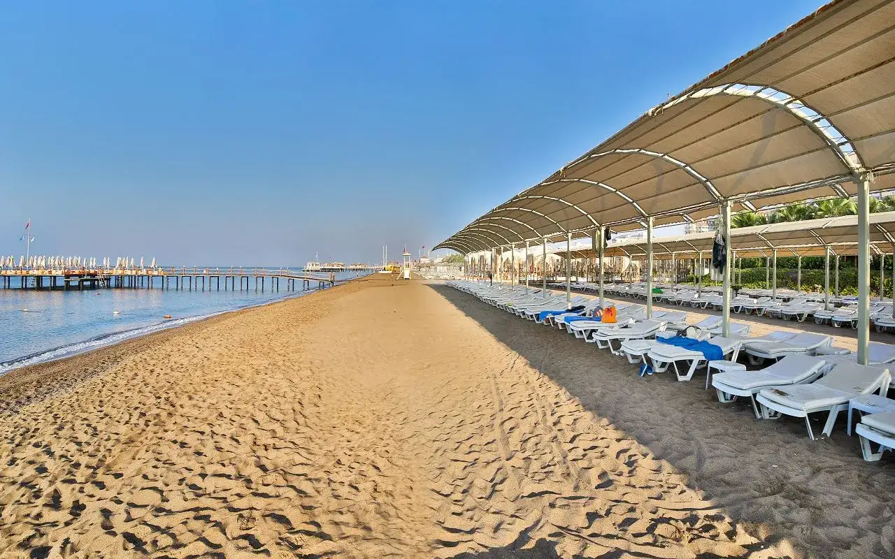 Pusta plaża w Lara, Turcja, z rzędami leżaków pod zadaszeniem i długim molo w oddali.