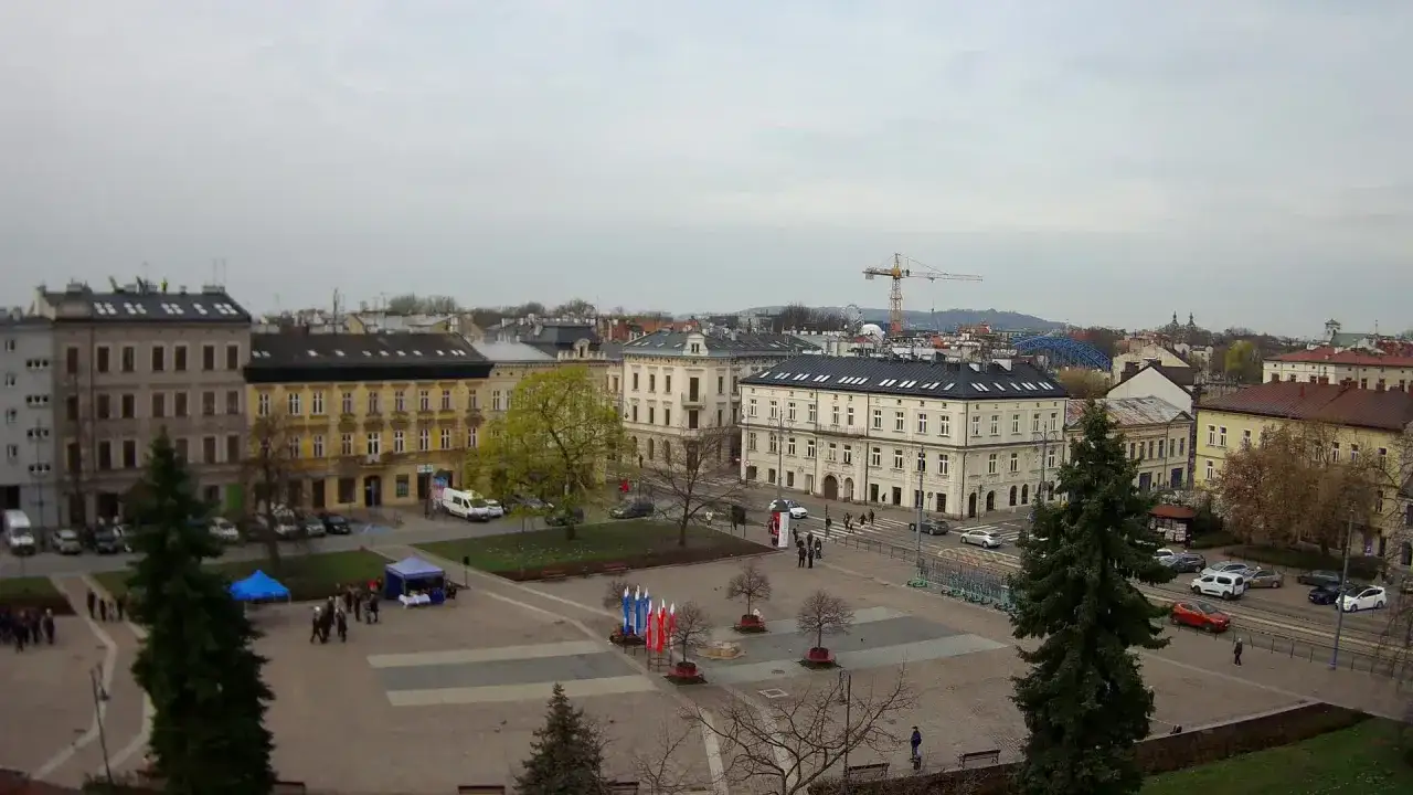 Widok na rynek podgórski z kamienicami, placem, drzewami i żurawiem budowlanym w tle.