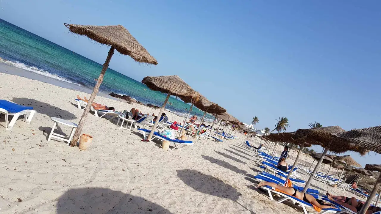 Djerba pogoda kwiecień: słoneczny dzień na plaży, leżaki pod strzechą, turkusowe morze, odpoczywający ludzie.