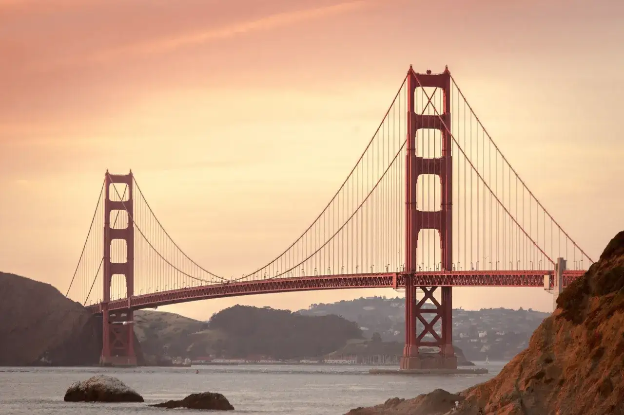 Ikoniczny most w San Francisco w ciepłych barwach zachodzącego słońca. Majestatyczna konstrukcja wznosi się nad wodą, otoczona wzgórzami.
