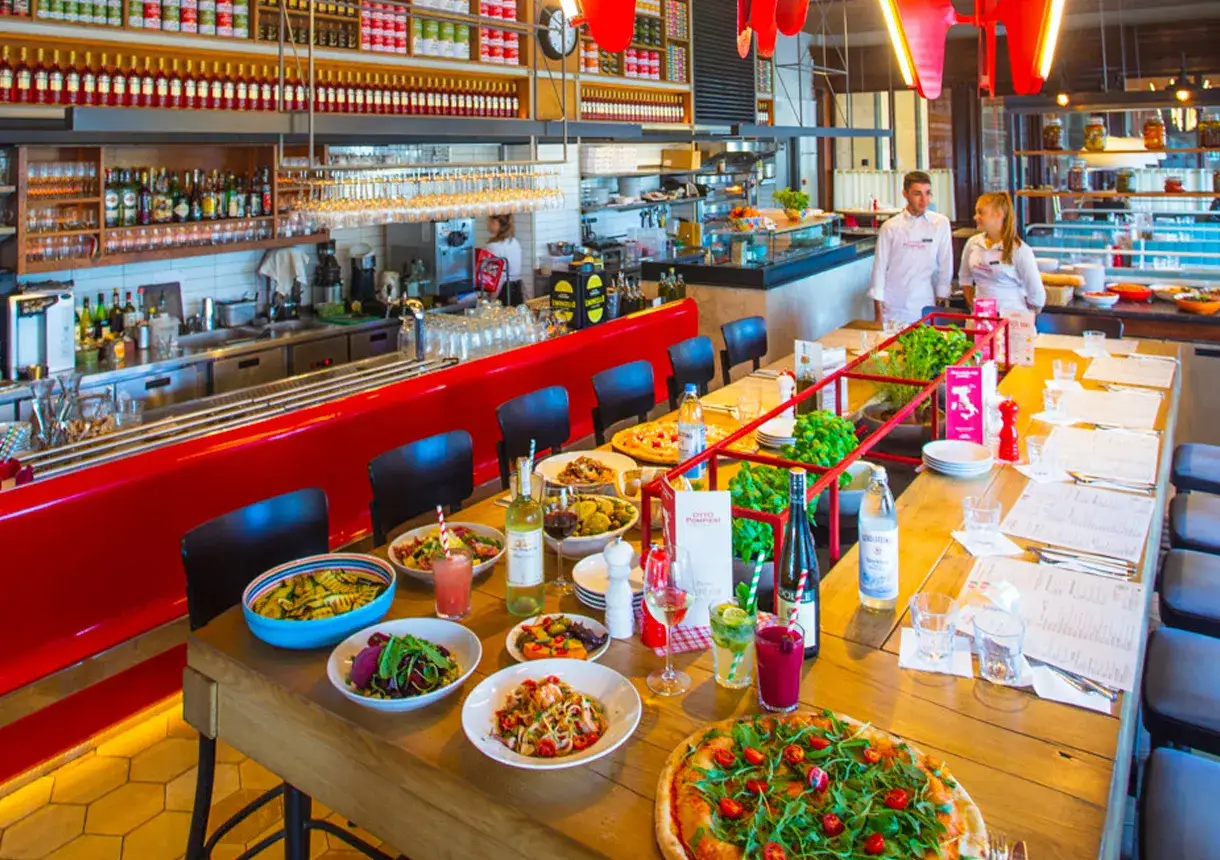 Stół w restauracji Otto Pompieri pełen pysznych dań, od pizzy po sałatki. W tle widać personel i bogato zaopatrzony bar.