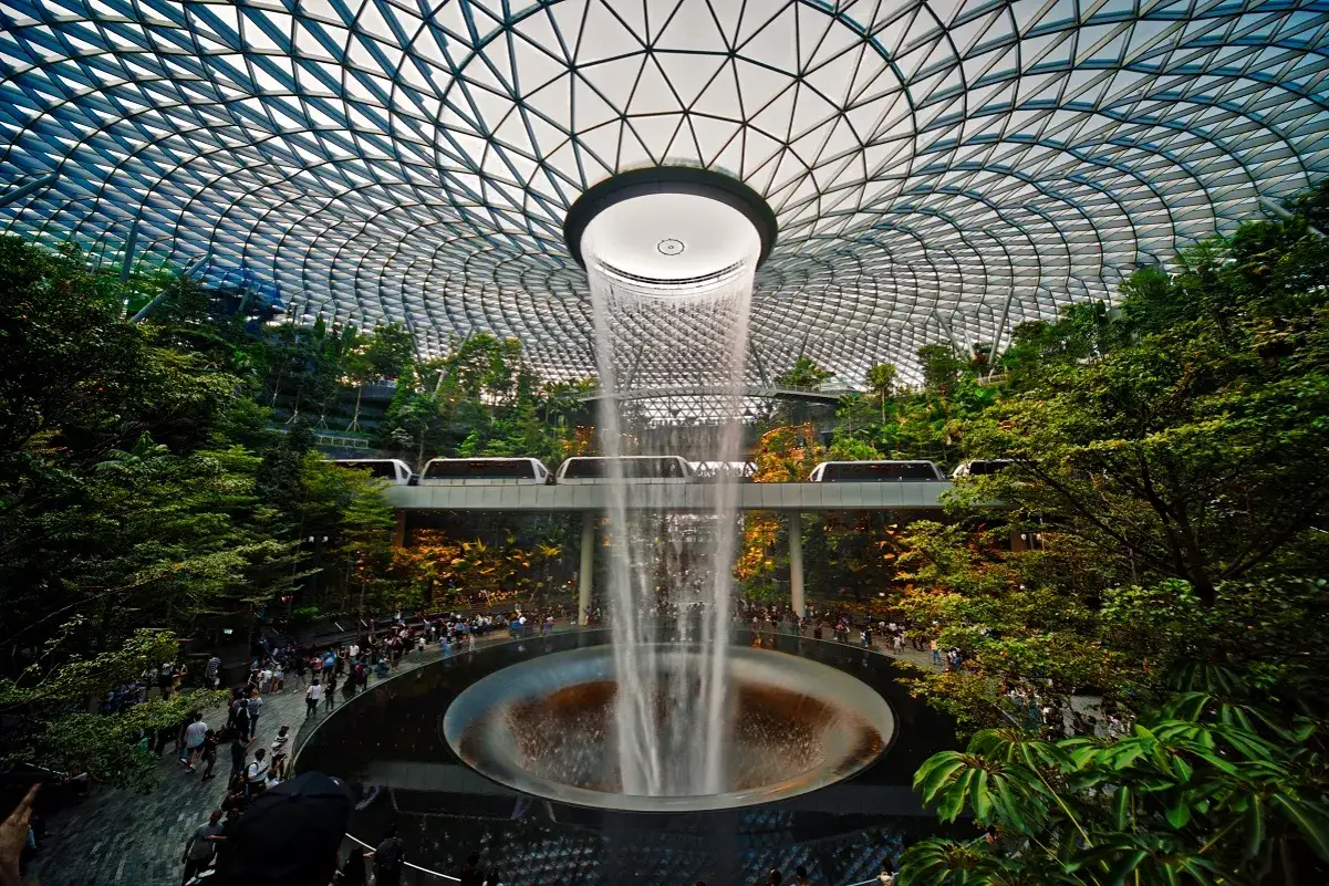Wodospad HSBC Rain Vortex w centrum handlowym Jewel Changi Airport w Singapurze, z kolejką naziemną przejeżdżającą nad zielonymi tarasami.