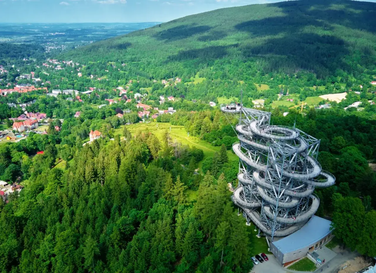 Widok na wieżę widokową i zielone góry w Polsce. Spiralne schody prowadzą na szczyt, skąd rozpościera się panorama na lasy i miasteczko.
