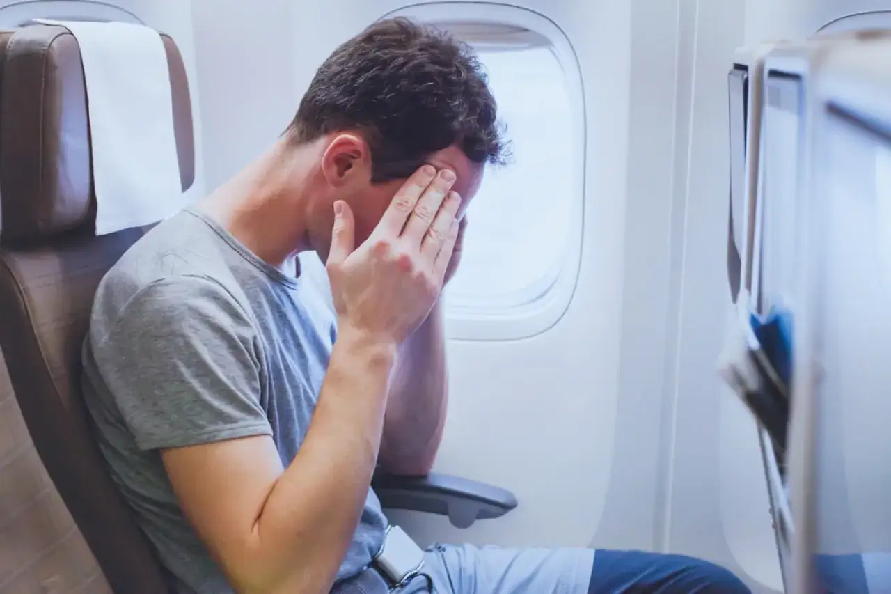 Mężczyzna w samolocie, z rękami na skroniach, wyraźnie cierpi na jetlag.