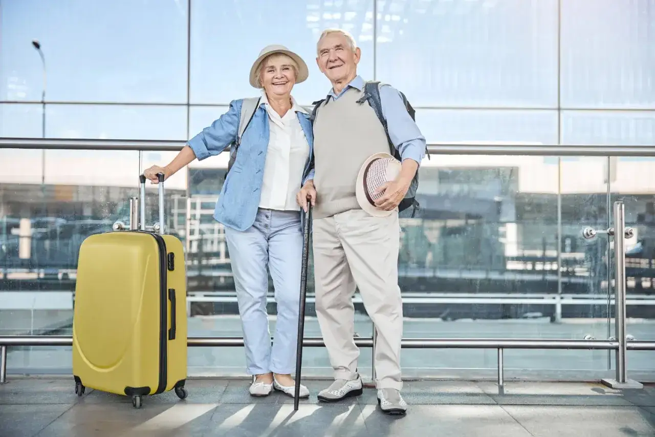 Uśmiechnięta para seniorów z walizką i laską, gotowa na wycieczki dla seniorów samolotem.