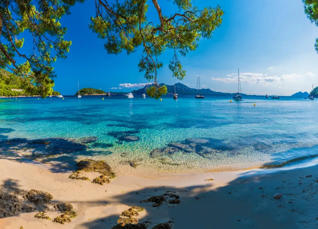 Piękna plaża z turkusową wodą i łodziami na horyzoncie. Idealne miejsce na wakacje na Balearach, jakie wyspy wybrać?
