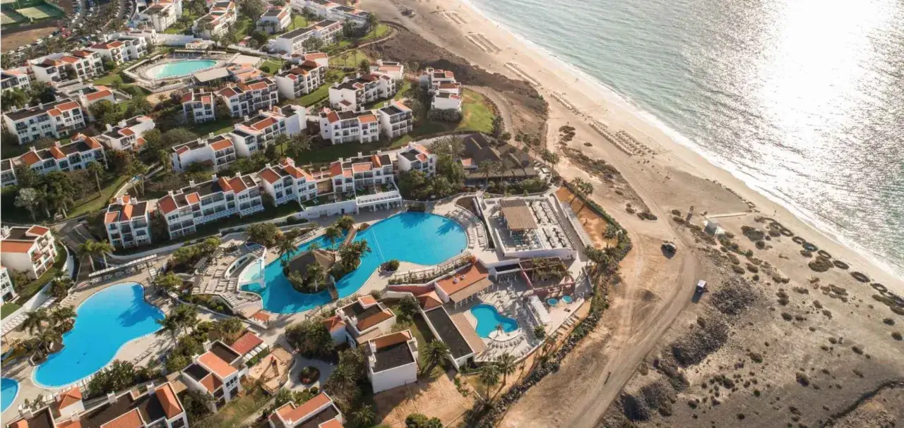 Widok z lotu ptaka na kompleks hotelowy z basenami i plażą. Wiele budynków z czerwonymi dachami. Fuerteventura Princess opinie potwierdzają urok tego miejsca.