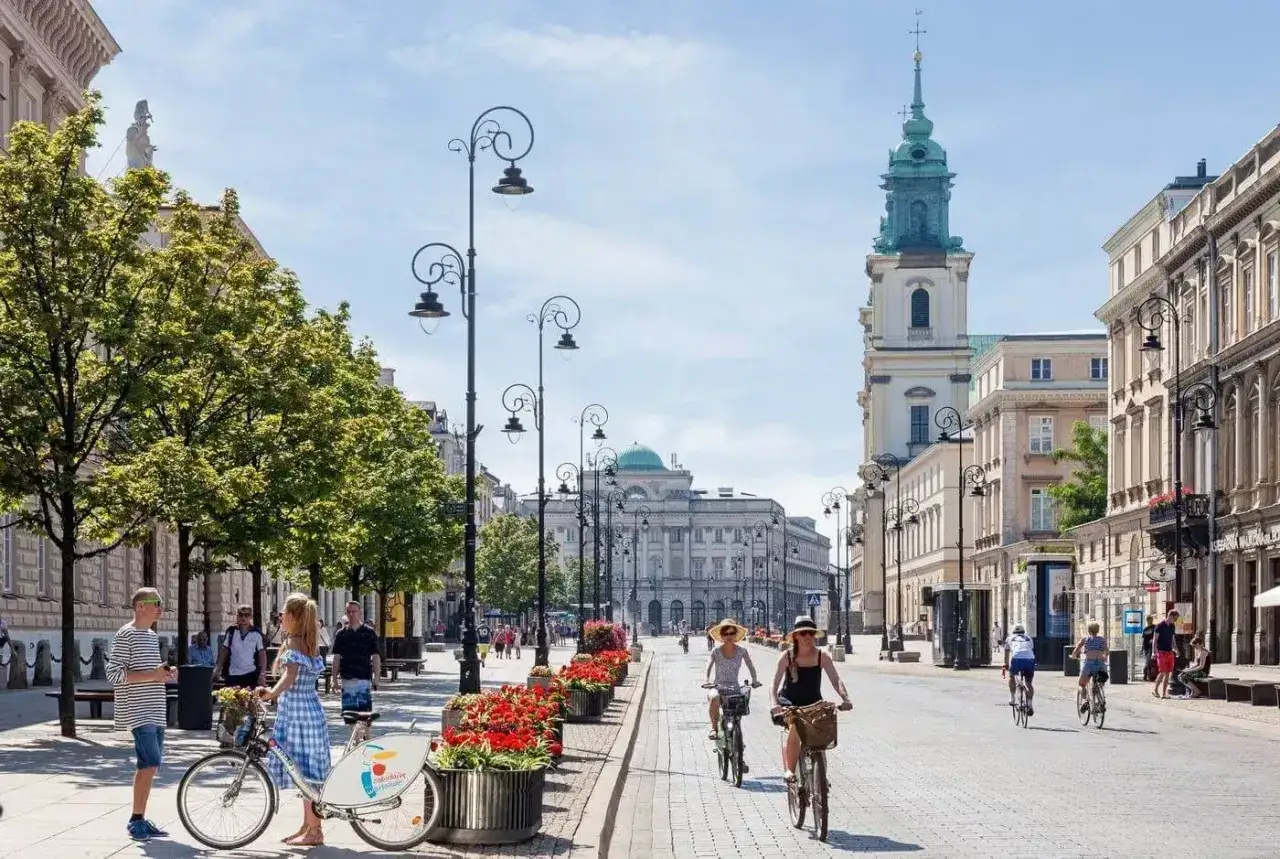 Słoneczny dzień na Krakowskim Przedmieściu. Ludzie spacerują i jeżdżą na rowerach wśród zieleni i zabytkowych kamienic.