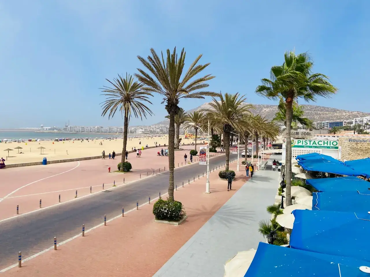 Agadir pogoda marzec: słoneczny dzień na plaży, palmy, spacerowicze i kawiarnie z niebieskimi parasolami.