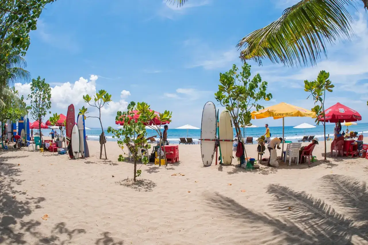 Słoneczna plaża w Kuta, deski surfingowe czekają na fale, parasole i palmy tworzą wakacyjny klimat.