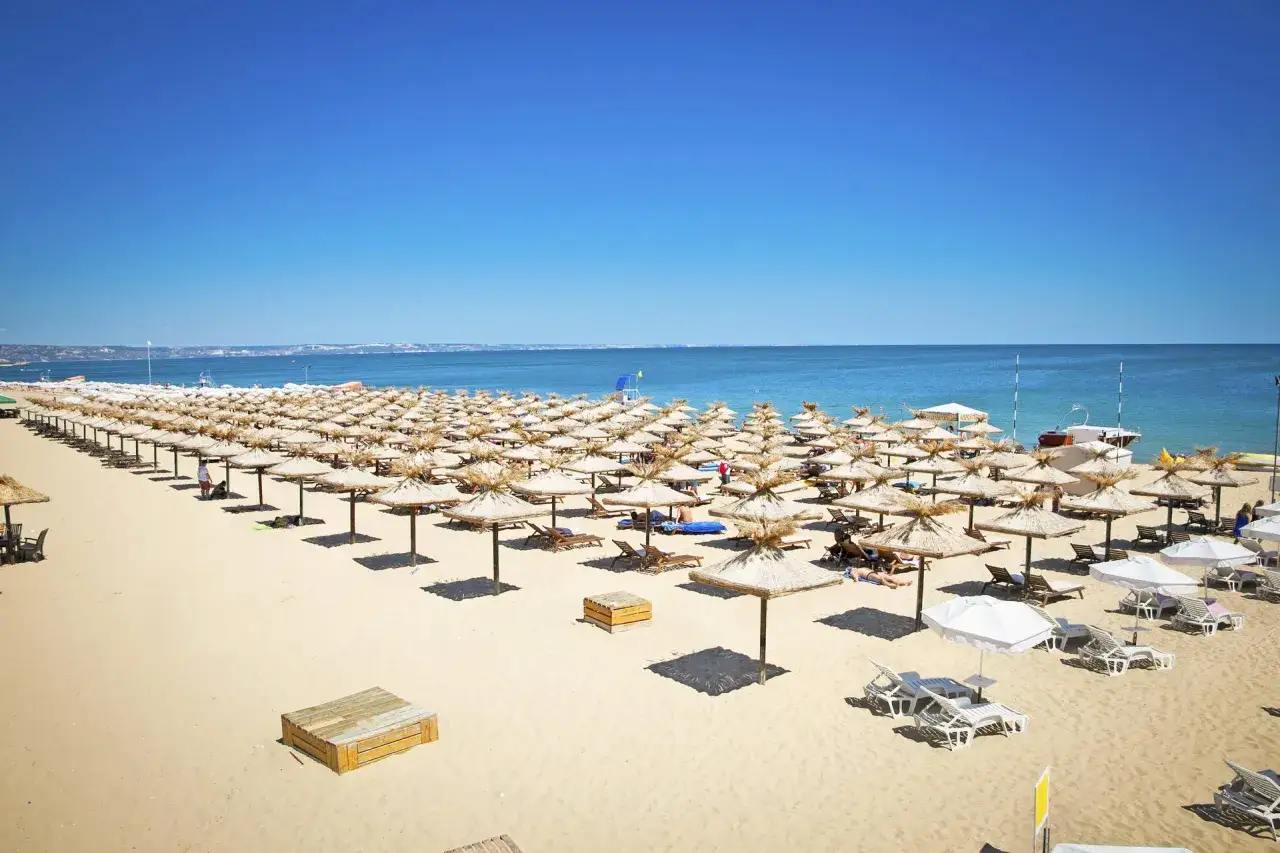Słoneczny brzeg w pełnym słońcu, z rzędami parasoli i leżaków czekających na plażowiczów.