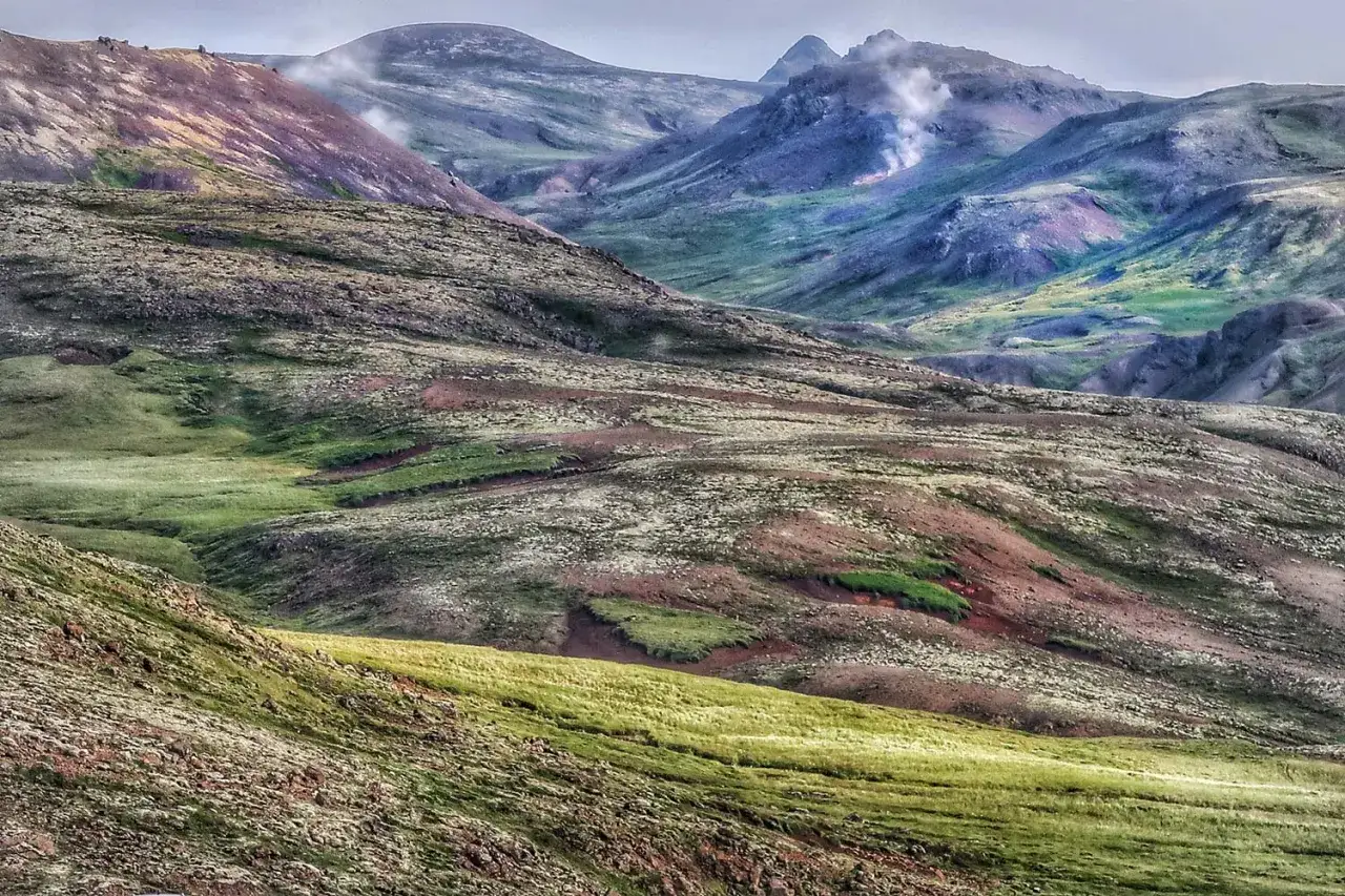 Górzyste krajobrazy Islandii, gdzie zieleń przeplata się z brązem i fioletem. W oddali widać parujące źródła geotermalne, tworzące złoty krąg na tle surowej przyrody.