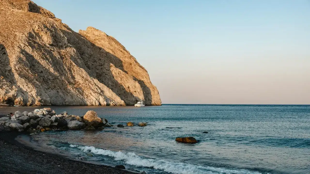 Skały i ciemny piasek na Santorini plażach, łódź kołysze się na spokojnym morzu przy skalistym wybrzeżu.