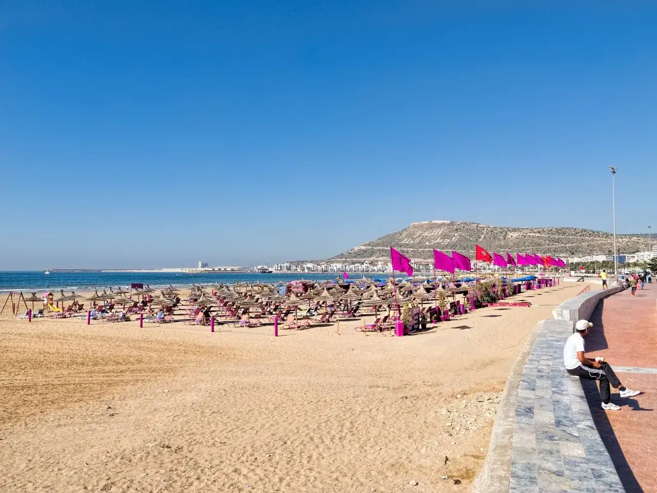 Agadir pogoda marzec: słoneczna plaża z leżakami i parasolami, w tle miasto i wzgórze z napisem.