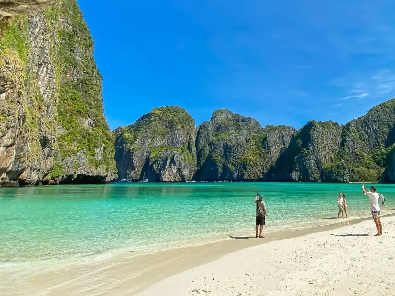 Rajska plaża Maya Bay z turkusową wodą i skalistymi klifami. Kilka osób spaceruje po piasku, podziwiając widok.