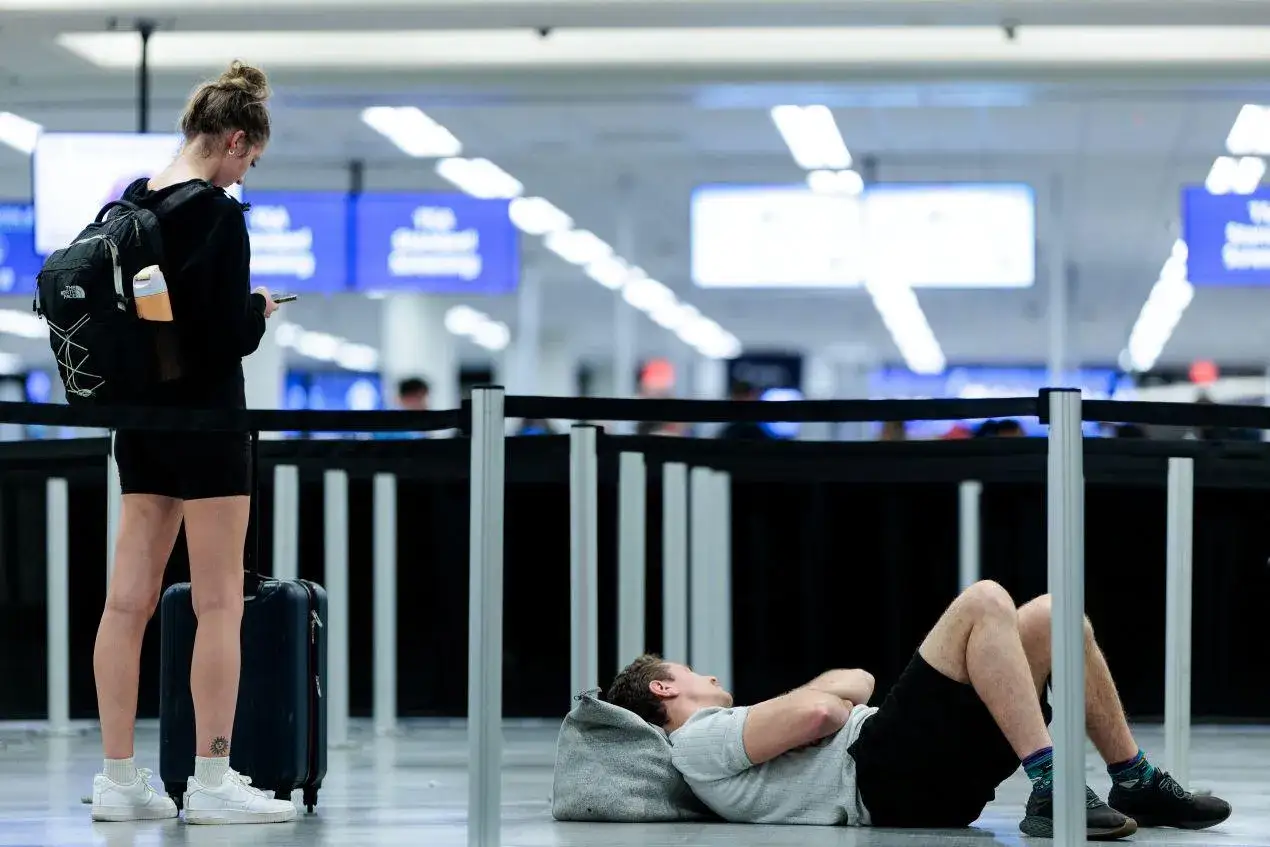 Zmęczona podróżna z plecakiem i walizką czeka na lot, podczas gdy mężczyzna śpi na podłodze. Szukając najlepszych reisen opinie, warto sprawdzić, czy linie lotnicze zapewniają komfort.
