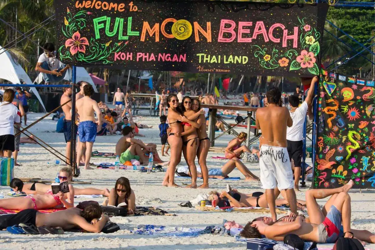 Plaża Koh Phangan, Tajlandia, przygotowuje się na full moon party. Ludzie odpoczywają na piasku, a nad nimi wisi kolorowy baner.