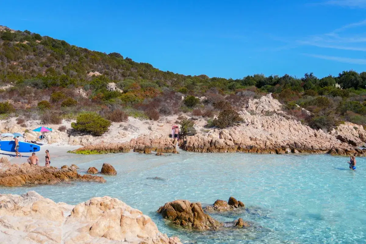 Słoneczna plaża na Costa Smeralda z turkusową wodą, skałami i ludźmi odpoczywającymi na piasku.
