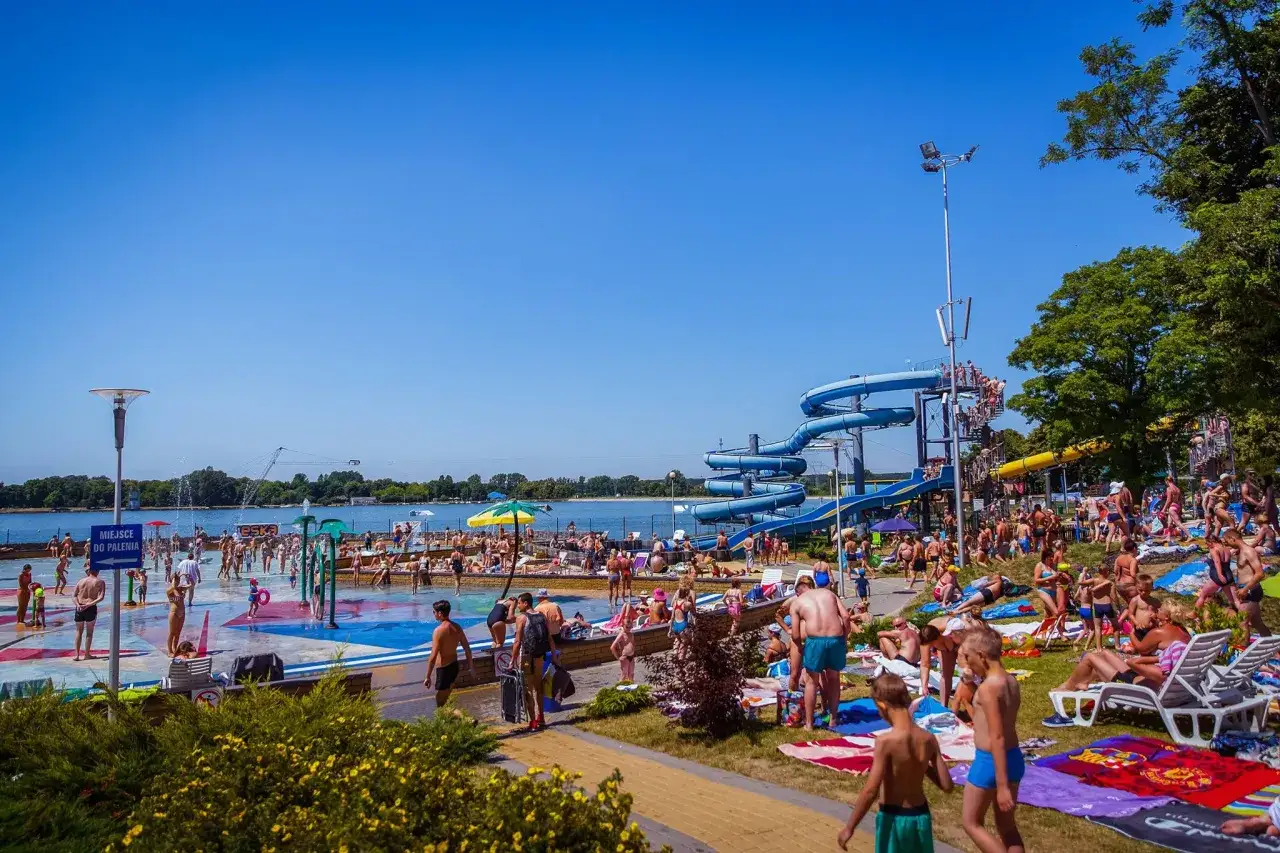 Tłumy ludzi korzystają z atrakcji wodnych na zalewie zemborzyckim. Widać zjeżdżalnie, brodziki i plażowiczów odpoczywających na leżakach.