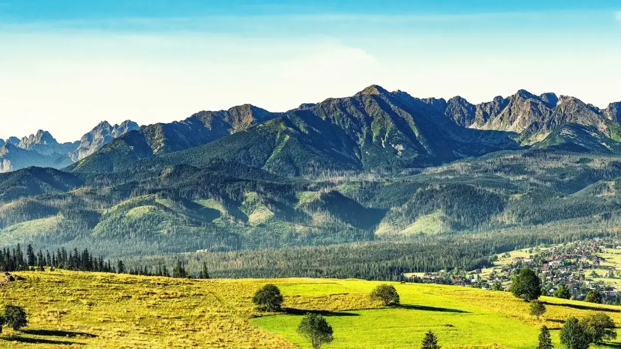 Widok na Tatry z Zakopanego. Zielone łąki i lasy schodzą ku malowniczej wsi, a w tle piętrzą się majestatyczne zakopane góry.