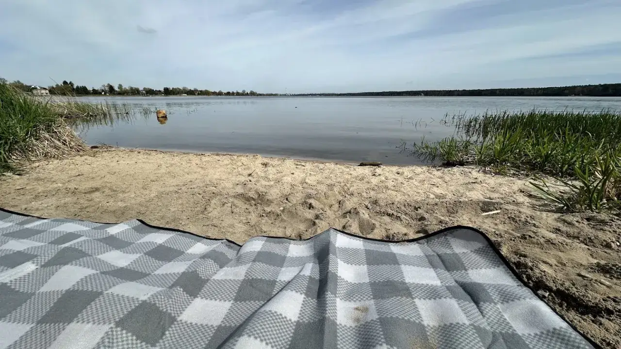 Piknik nad zalewem zemborzyckim. Piaszczysty brzeg, woda i zielona trawa. Koc w kratę czeka na gości.