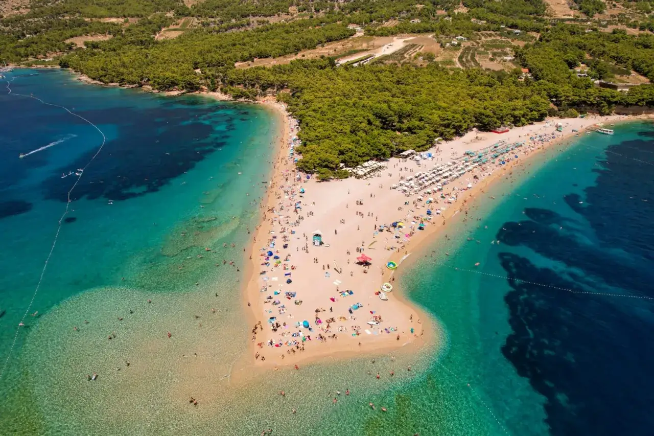 Idealne miejsce, gdzie na wakacje w sierpniu: piaszczysta plaża otoczona turkusowym morzem i zielonym lasem, pełna wypoczywających ludzi.