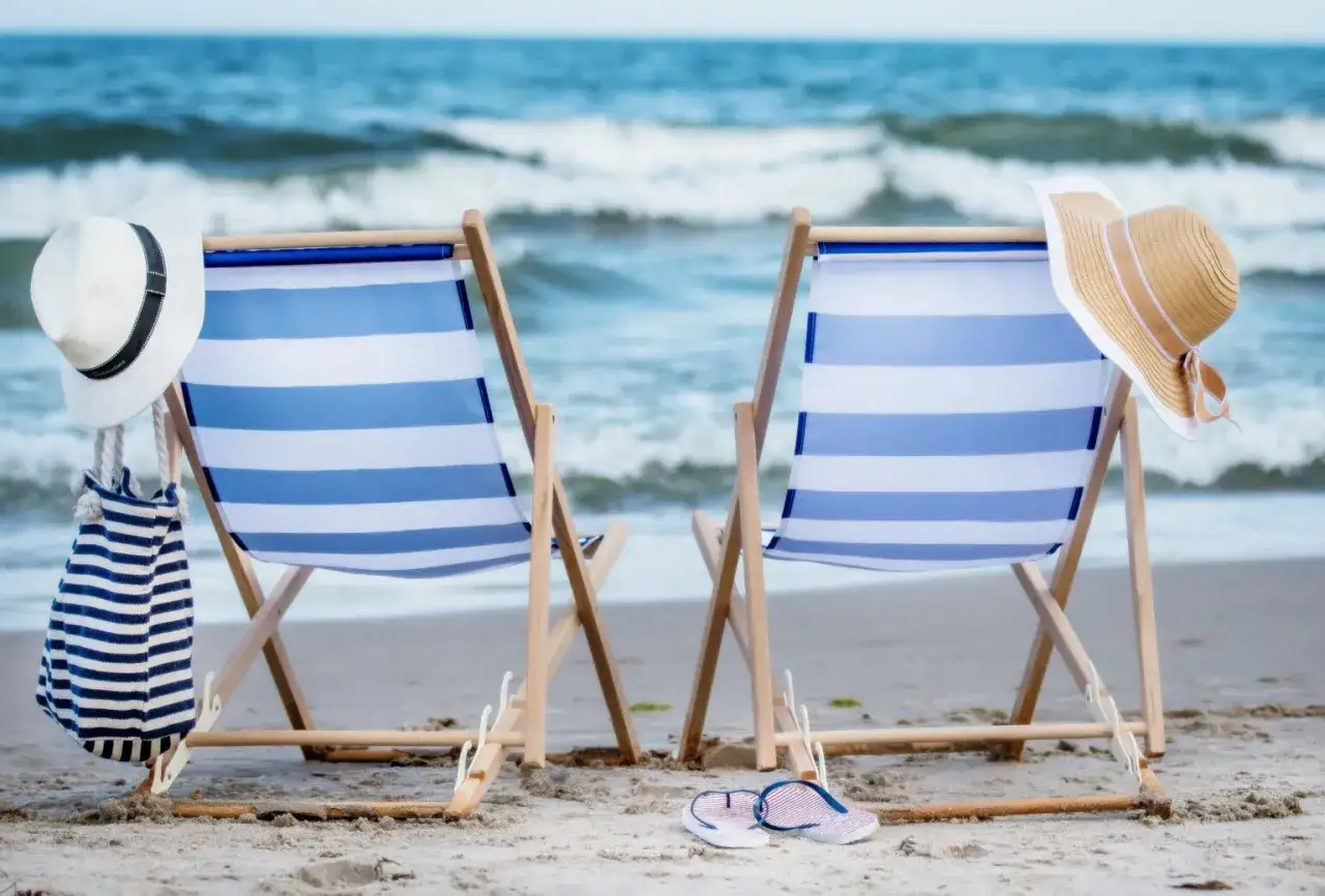 Dwie leżaki na plaży, gotowe na 10 dniowe wczasy nad morzem z wyżywieniem. W tle fale, na leżakach kapelusze i torba.