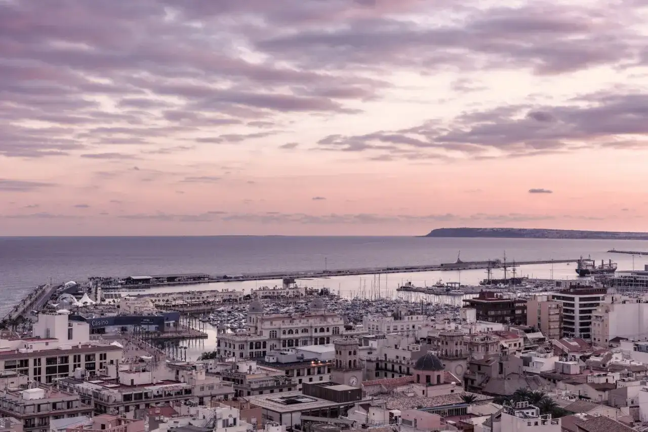 Alicante, grudzień. Zachód słońca nad portem z jachtami i budynkami. Pogoda łagodna, niebo w odcieniach różu i fioletu.