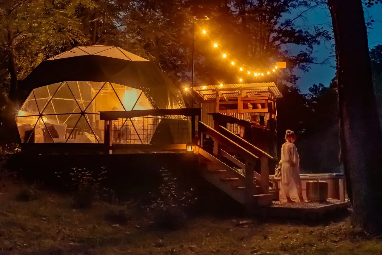 Nocą, przy blasku girlandy świateł, rozświetlona kopuła obiecuje romantyczny weekend dla dwojga tanio. Obok, drewniana konstrukcja z dachem.