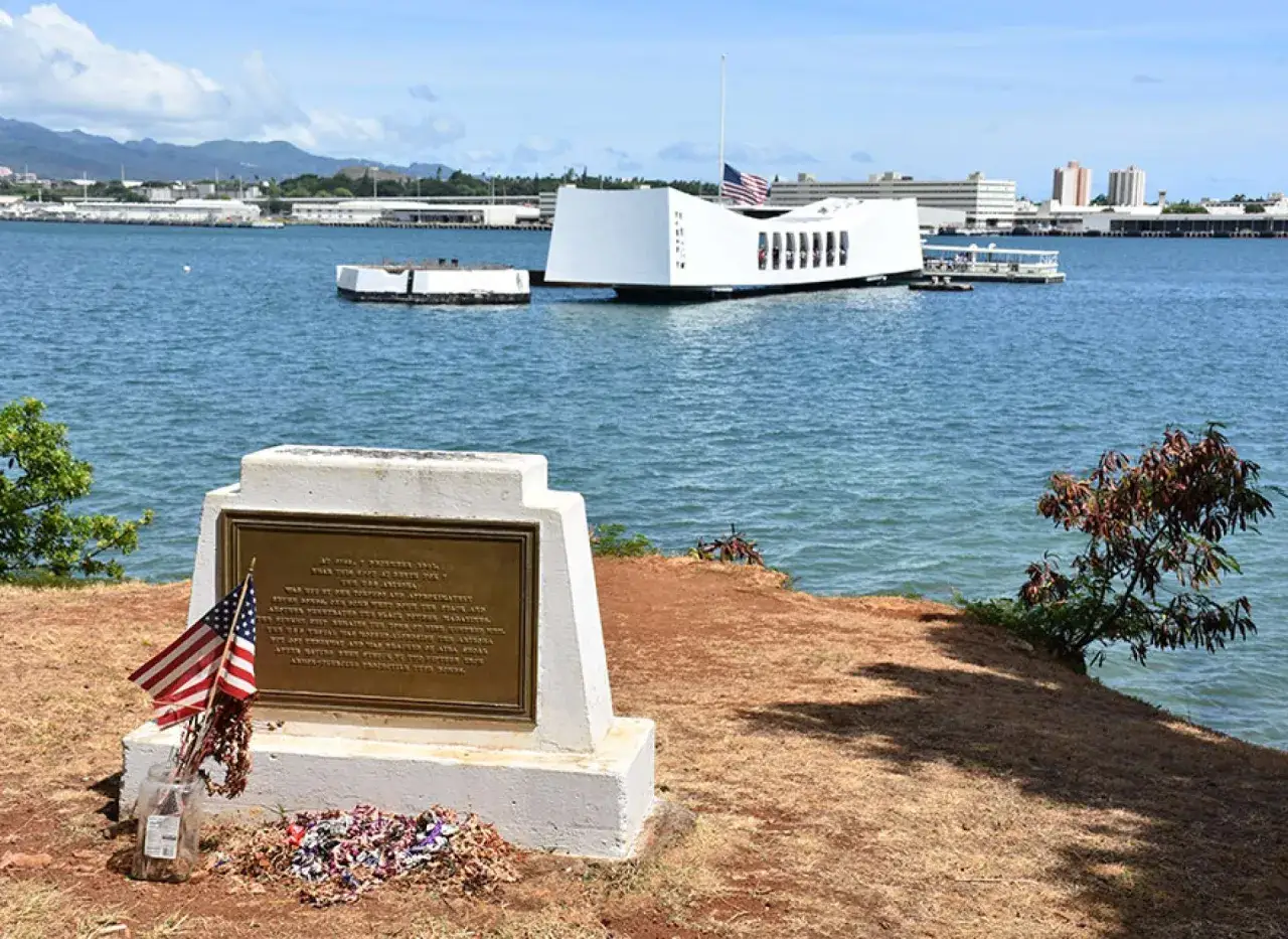 Pomnik upamiętniający atak na Pearl Harbor. Na pierwszym planie tablica z flagami USA i wieńcem. W tle biała konstrukcja memoriału nad wodą.