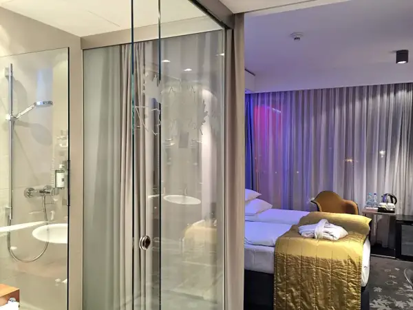 Łazienka i sypialnia w hotelu Puro Wrocław. Szklane drzwi oddzielają prysznic od pokoju z łóżkiem i zasłonami.