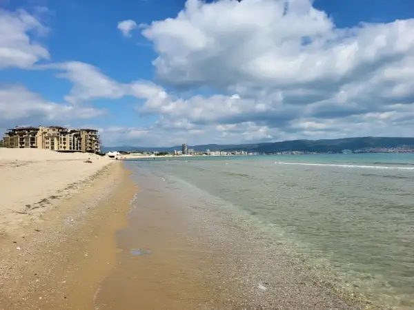 Słoneczny brzeg z piaszczystą plażą, turkusową wodą i hotelami w tle. Chmury na błękitnym niebie zapowiadają piękną pogodę.