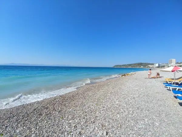 Słoneczna plaża w Ixia, Rodos, z turkusowym morzem i kamienistym brzegiem. Dwie osoby odpoczywają pod parasolem.