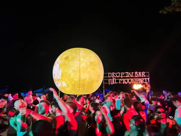 Tłum ludzi bawi się na plaży podczas **full moon party**. W tle neonowy napis "DROP IN BAR FULL MOON PARTY" i wielka, świecąca kula imitująca księżyc.