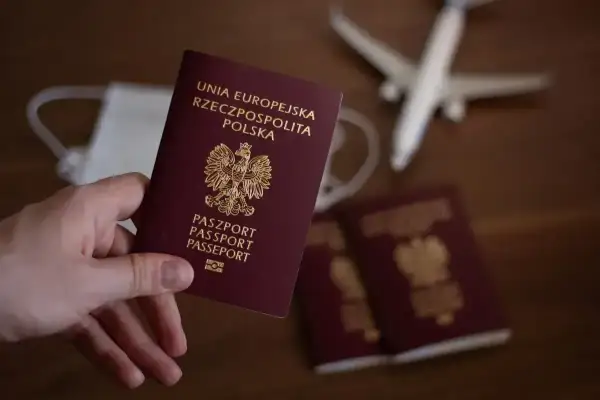 Dłoń trzyma polski paszport. Obok leżą kolejne paszporty, maseczka i samolot, sugerując podróż i pytając, ile wyrabia się paszport.