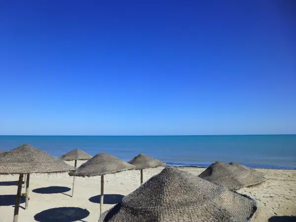 Słoneczna plaża na Djerbie w kwietniu. Puste parasole z trawy na piasku, turkusowe morze i bezchmurne niebo.