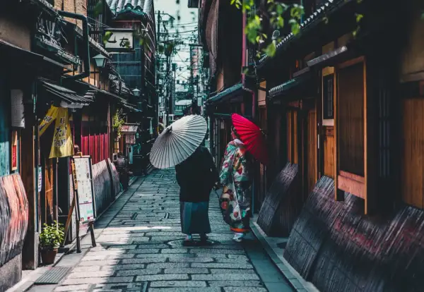 Dwie kobiety w kimonach spacerują wąską uliczką w Kioto, trzymając parasolki. To fascynujące ciekawostki o Japonii.
