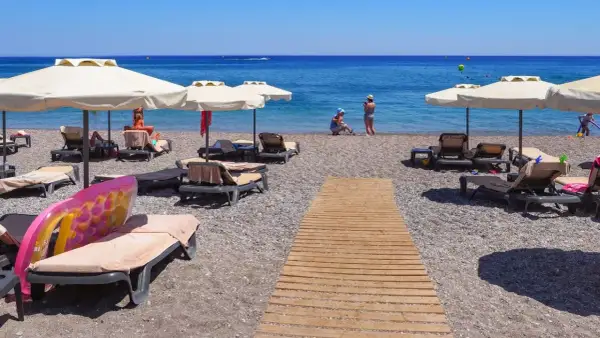 Słoneczny kurort nad Morzem Czarnym. Leżaki pod parasolami, drewniana ścieżka na kamienistej plaży, ludzie kąpiący się w morzu.